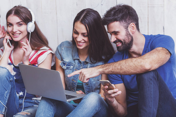 Group of attractive young people sitting on the floor using a laptop, Tablet PC, smart phones, headphones listening to music, smiling - Φωτογραφία, εικόνα