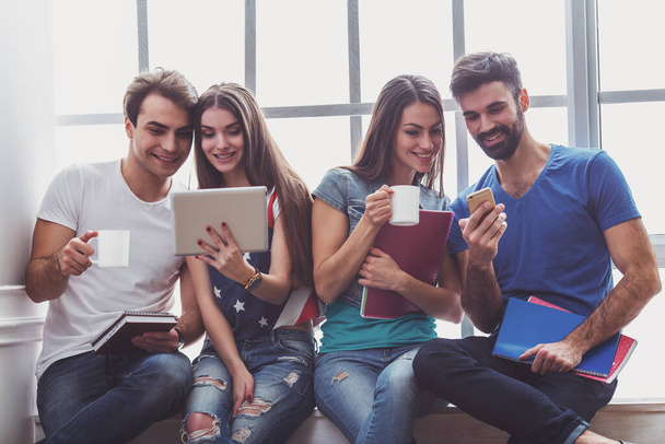 Quatro jovens atraentes, mantendo à mão computador tablet, copo, smartphone e notebook, sentar-se no peitoril da janela, sorrindo
 - Foto, Imagem