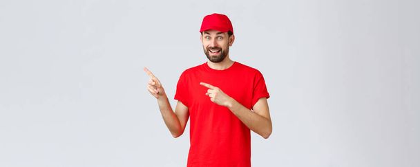 Online shopping, delivery during quarantine and takeaway concept. Amused and excited smiling courier inviting client take look, employee in red uniform cap and t-shirt pointing fingers left - Photo, Image