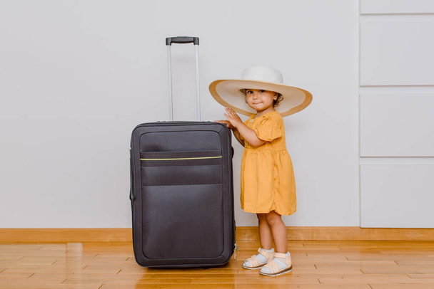 A little girl in white oversize hat near a big suitcase. Children want in a vacation concept - Zdjęcie, obraz