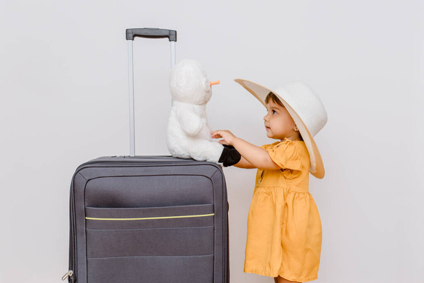 A little girl in white oversize hat near a big suitcase is playing with her plush snowman toy. Children want in a vacation concep - Фото, изображение