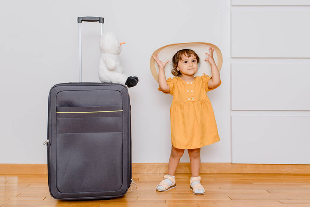 A little girl in white oversize hat near a big suitcase is playing with her plush snowman toy. Children want in a vacation concep - 写真・画像
