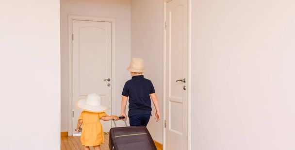 Back view of a happy girl and boy, brother and sister  are carrying a big suitcase in room with white walls and wood floor, and expecting the big vacation - Фото, изображение