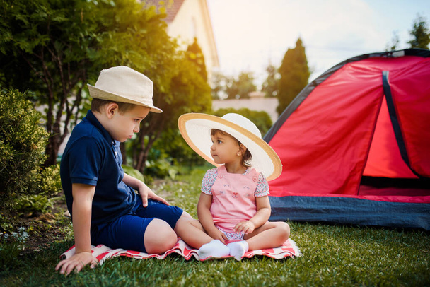 Two happy little kids, boy and girl brother and sister are siting and playing on the grass near a red camping tent  in the home yard. Home camping vacation concept - Zdjęcie, obraz