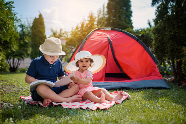 İki mutlu çocuk, erkek ve kız, erkek ve kız kardeş ve kız kardeş çimlerin üzerinde oturuyorlar ve erkek kardeş, evlerinin bahçesindeki kırmızı bir kamp çadırının yanında kız kardeşine peri masalları okuyor. Ev kampı tatil konsepti - Fotoğraf, Görsel