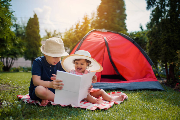 Two happy little kids, boy and girl, brother and sister are sitting on the grass and brother is reading fairy tales to his sister near a red camping tent in their home yard. Home camping vacation concept - Photo, image