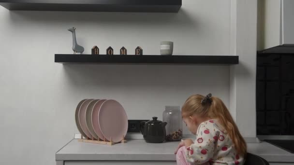 a little blonde girl in pajamas has breakfast in a bright kitchen. High quality FullHD footage - Materiał filmowy, wideo
