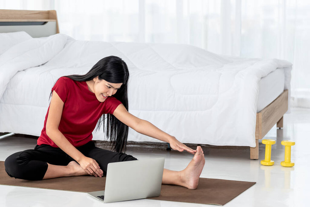Mujer asiática, atractiva y joven, está practicando yoga estudiando clases en línea, en su dormitorio blanco, concepto de nueva normalidad y educación en línea - Foto, Imagen