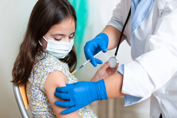 Little caucasian girl with face mask watching her being vaccinated by a medical worker. Child vaccination concept. - Photo, image