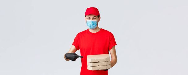 Food delivery, application, online grocery, contactless shopping and covid-19 concept. Cheerful courier in red uniform, face mask and gloves pointing finger at pizza boxes, deliver to client house - Photo, Image