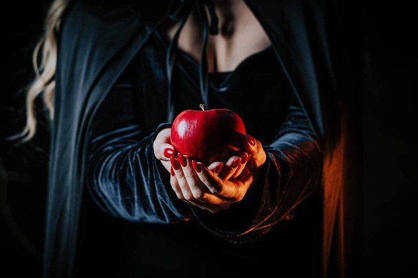 Witch woman in black offers red apple as symbol of temptation, poison. Fairy tale, white snow wizard concept. Spooky halloween, cosplay.  - Фото, изображение
