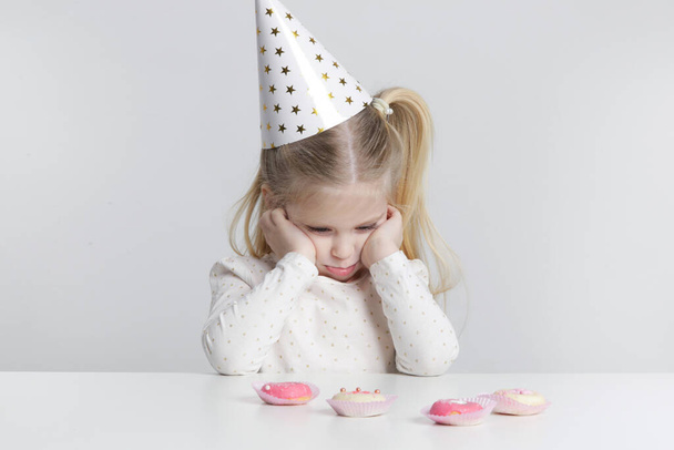 Displeased little girl with birthday hat and donuts in front of her. - 写真・画像