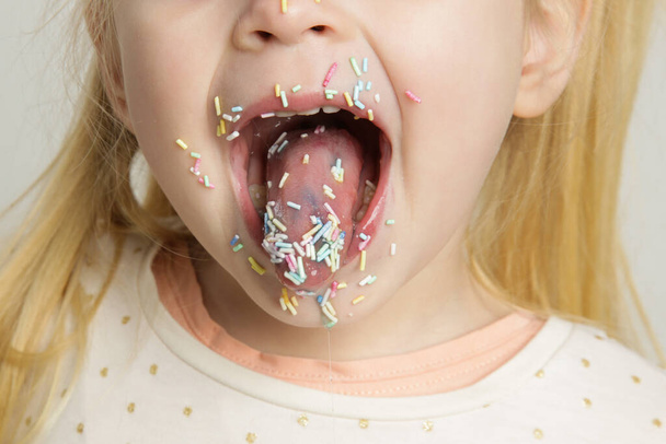 Cute little birthday girl with colorful sugar sprinkles on lips and in mouth. Children and sugar addiction concept. - 写真・画像