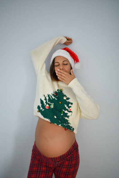 A young pregnant Caucasian female with a Santa hat and a sweater with a Christmas tree - 写真・画像