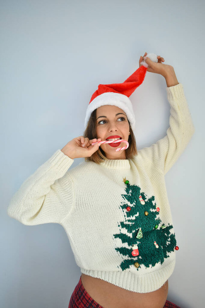 A young pregnant Caucasian female with a Santa hat eating a candy cane - Fotó, kép