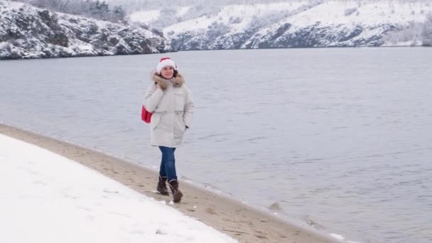 Жінка в капелюсі Сантаса з червоною сумкою йде вздовж річки взимку Різдва
 - Кадри, відео