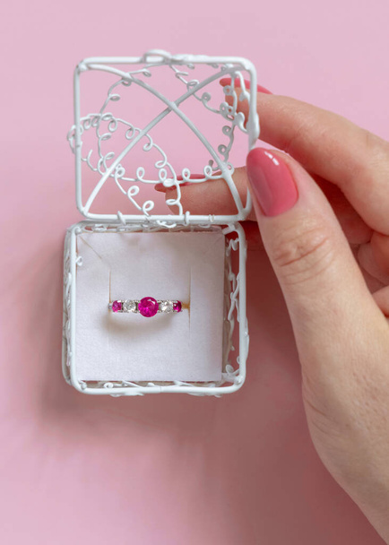 Жіноча рука відкриває прикрашену подарункову коробку з кільцем втягування дорогоцінного каміння на рожевий вид зверху. Концепція шлюбної пропозиції, сцена дня валентинки
 - Фото, зображення