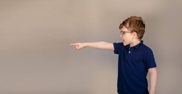 Kleine jongen in blauw t-shirt en bril op grijze achtergrond wijzen met zijn vinger en hebben een boze uitdrukking op zijn gezicht. Vrije ruimte voor tekst en logo - Foto, afbeelding