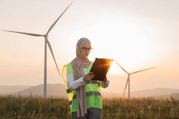 Woman in hijab standing on windmill farm with clipboard - Foto, Imagem