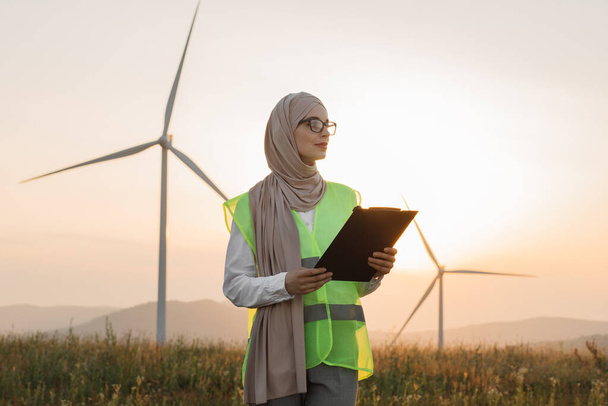 Muslim woman holding clipboard while posing on windmill farm - Foto, afbeelding