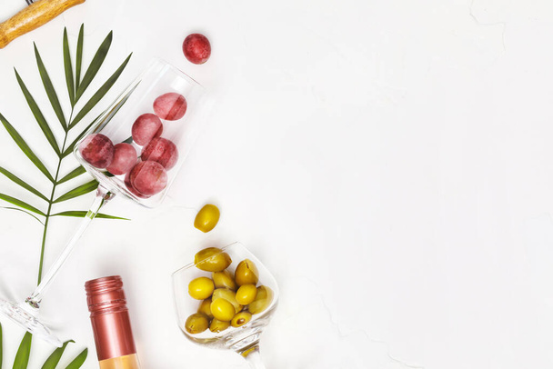 Composition gastronomique méditerranéenne avec verres à vin avec raisins et olives vertes, bouteille de vin de rose et feuilles de palmier sur fond blanc avec espace de copie. Le concept d'une station de vacances d'été et se détendre - Photo, image
