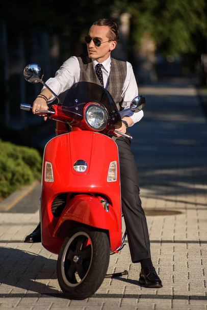 Młody przystojny mężczyzna w eleganckich klasycznych ubraniach siedzi na czerwonym vintage skuterze motocyklowym na ulicy - Zdjęcie, obraz