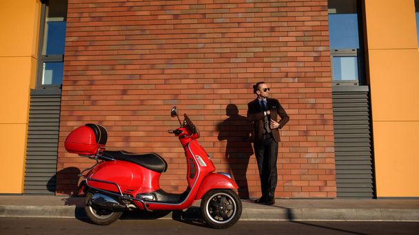 Man in klassieke pak in de buurt van een rode vintage scooter op een bakstenen muur - Foto, afbeelding