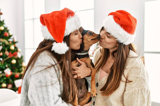 Deux femmes embrassant et étreignant chien debout près de l'arbre de Noël à la maison - Photo, image