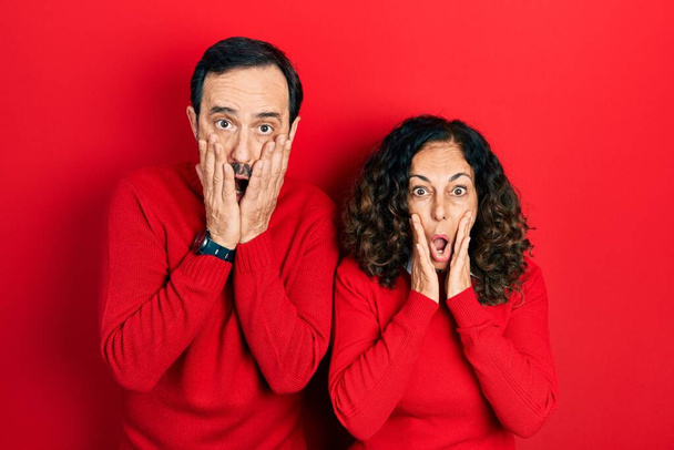 Middle age couple of hispanic woman and man hugging and standing together afraid and shocked, surprise and amazed expression with hands on face  - 写真・画像
