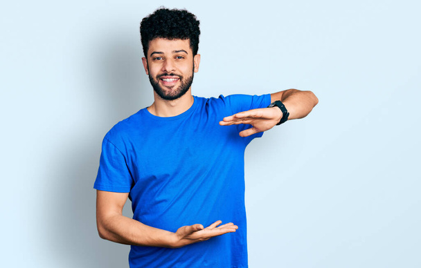 Young arab man with beard wearing casual blue t shirt gesturing with hands showing big and large size sign, measure symbol. smiling looking at the camera. measuring concept.  - Фото, изображение