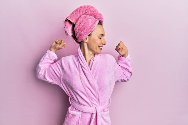 Young blonde woman wearing shower towel cap and bathrobe stretching back, tired and relaxed, sleepy and yawning for early morning  - Photo, image