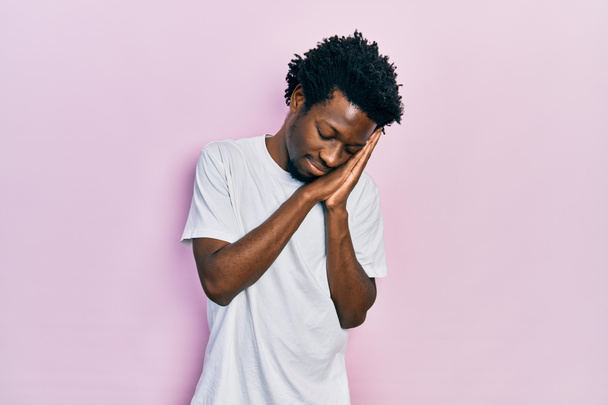 Young african american man wearing casual white t shirt sleeping tired dreaming and posing with hands together while smiling with closed eyes.  - Foto, Imagem