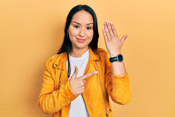 Beautiful hispanic woman with nose piercing wearing yellow leather jacket in hurry pointing to watch time, impatience, looking at the camera with relaxed expression  - Foto, afbeelding
