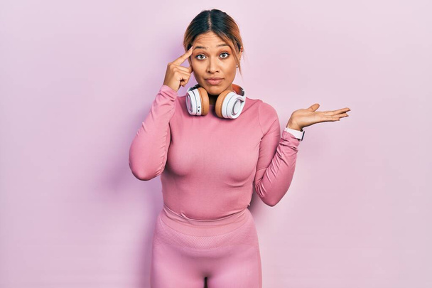 Beautiful hispanic woman wearing gym clothes and using headphones confused and annoyed with open palm showing copy space and pointing finger to forehead. think about it.  - Photo, Image