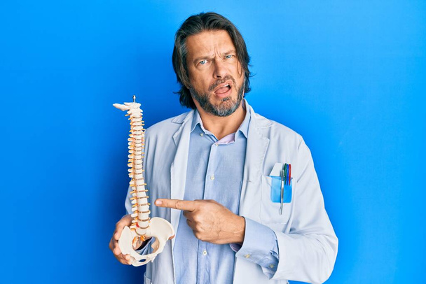 Middle age handsome traumatologist man holding anatomical model of spinal column in shock face, looking skeptical and sarcastic, surprised with open mouth  - Foto, Imagen