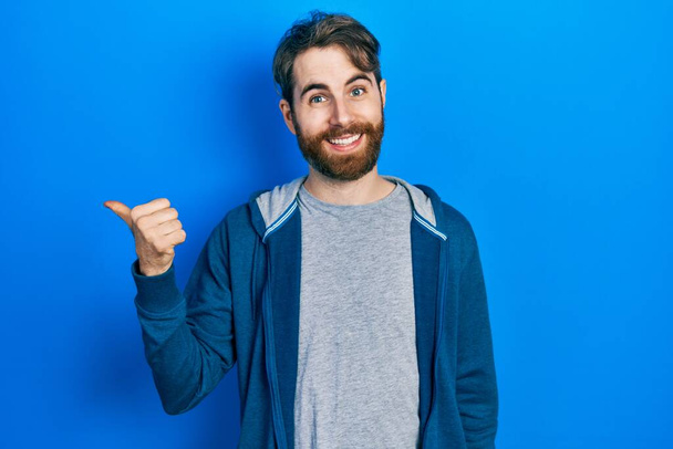 Caucasian man with beard wearing casual sweatshirt smiling with happy face looking and pointing to the side with thumb up.  - Photo, Image