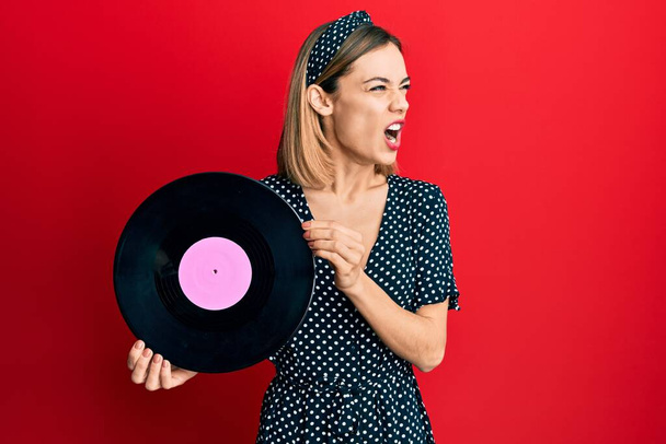 Young caucasian blonde woman holding vinyl disc angry and mad screaming frustrated and furious, shouting with anger. rage and aggressive concept.  - Photo, Image
