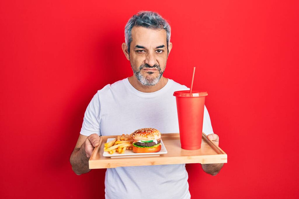 Handsome middle age man with grey hair eating a tasty classic burger with fries and soda skeptic and nervous, frowning upset because of problem. negative person.  - Valokuva, kuva