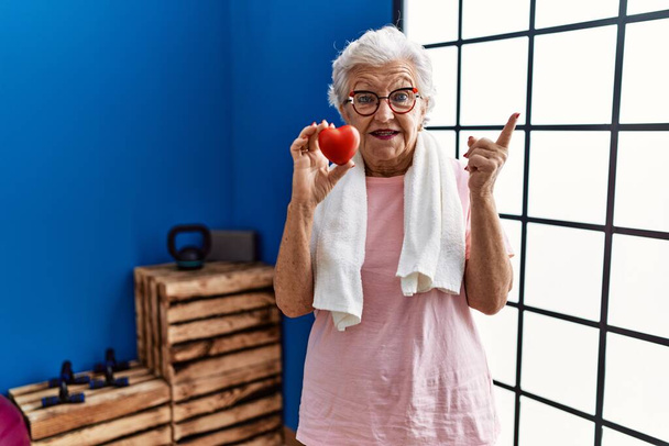 Старша жінка з сірим волоссям в спортивному одязі, що тримає червоне серце посміхаючись щасливо вказуючи рукою і пальцем на бік
  - Фото, зображення