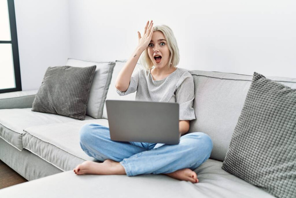 Young caucasian woman using laptop at home sitting on the sofa surprised with hand on head for mistake, remember error. forgot, bad memory concept.  - Foto, Bild