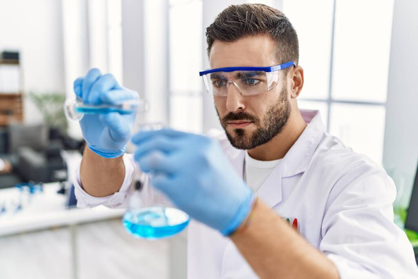 Junger hispanischer Mann in Wissenschaftleruniform mischt Flüssigkeit im Labor - Foto, Bild