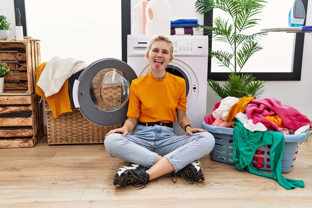 Young blonde woman doing laundry sitting by washing machine sticking tongue out happy with funny expression. emotion concept.  - Foto, afbeelding