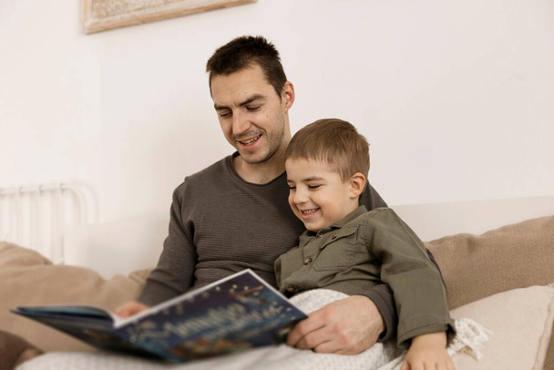Отец и сын читают книгу на кровати дома. Молодой привлекательный мужчина и маленький мальчик отдыхают в спальне. Естественные цвета земли. Уютная обстановка. Отец читает сказку для своего ребенка. - Фото, изображение