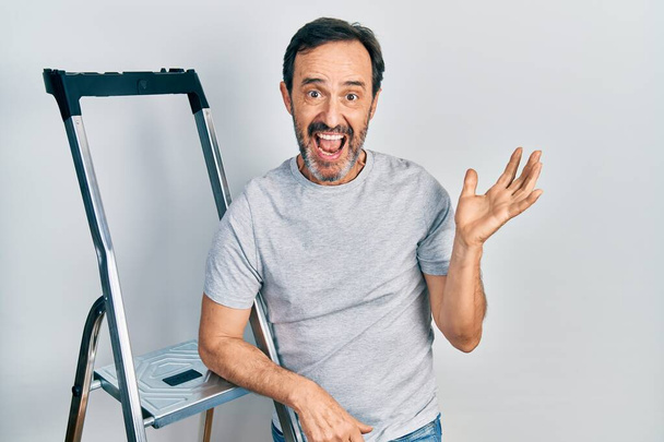 Middle age hispanic man using ladder celebrating victory with happy smile and winner expression with raised hands  - Foto, immagini