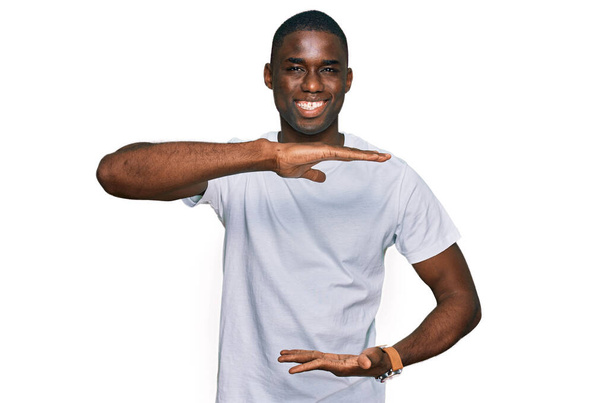 大小のサインを示す手でジェスチャーをカジュアルな白いTシャツを着て若いアフリカ系アメリカ人の男性は、シンボルを測定します。カメラを見て笑ってる。概念を測定し.  - 写真・画像