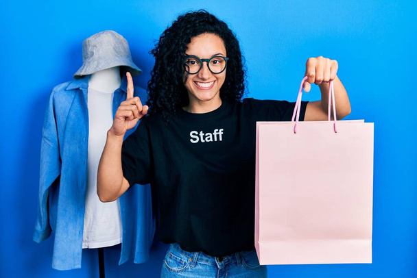 Νεαρή ισπανόφωνη γυναίκα με σγουρά μαλλιά φορώντας το μπλουζάκι του προσωπικού κρατώντας τσάντα για ψώνια χαμογελώντας με μια ιδέα ή ερώτηση δείχνοντας δάχτυλο με χαρούμενο πρόσωπο, νούμερο ένα  - Φωτογραφία, εικόνα