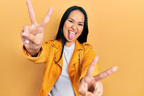 Beautiful hispanic woman with nose piercing wearing yellow leather jacket smiling with tongue out showing fingers of both hands doing victory sign. number two.  - Foto, afbeelding