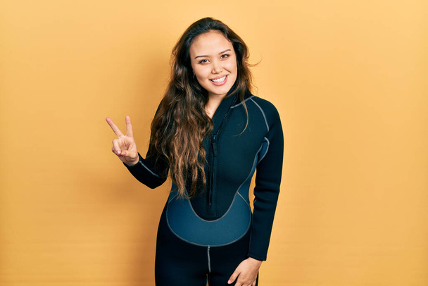 Young hispanic girl wearing diver neoprene uniform smiling looking to the camera showing fingers doing victory sign. number two.  - Valokuva, kuva