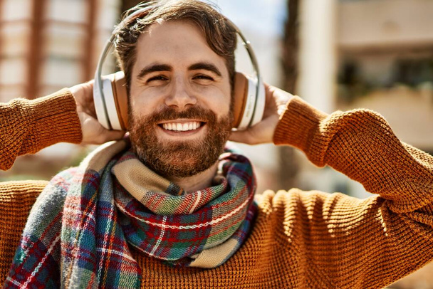 Jovem caucasiano com barba ouvindo música usando fones de ouvido ao ar livre em um dia ensolarado - Foto, Imagem