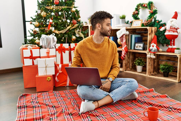 Arab young man using laptop sitting by christmas tree looking to side, relax profile pose with natural face and confident smile.  - Fotoğraf, Görsel